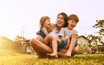A laughing mother sits cross-legged on grass at sunset, holding her two young children on her lap, all three laughing together, next to the Westfield Health logo.