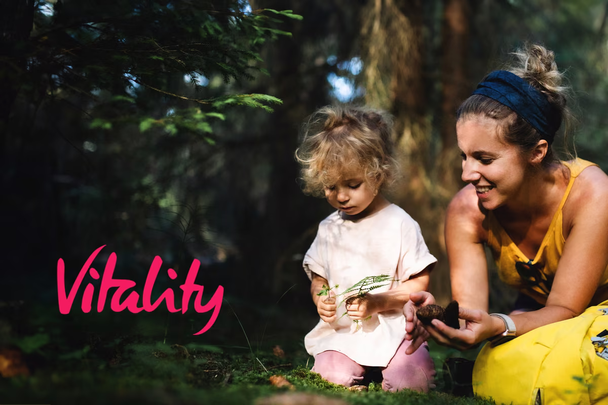 Woman and toddler examine a pine cone and fern together on forest grass. Vitality logo overlaid.