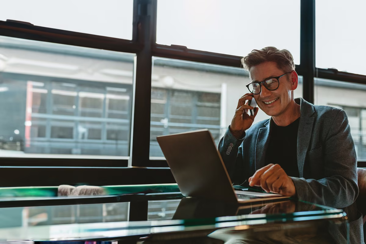 Businessman in a suit smiling on the phone while using a laptop at a glass desk