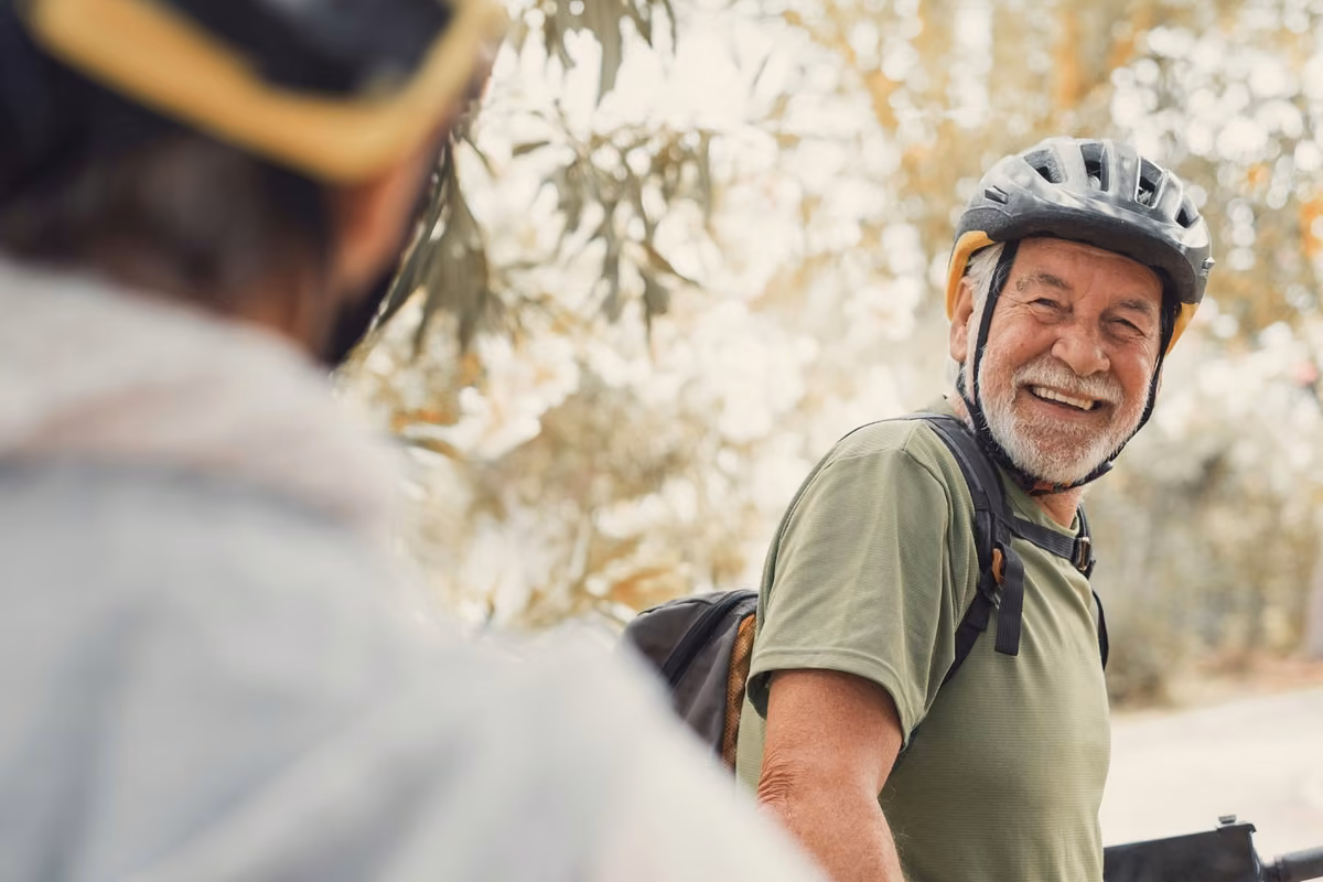 Older man in a cycling helmet and backpack smiles at a companion outdoors, with trees in the background