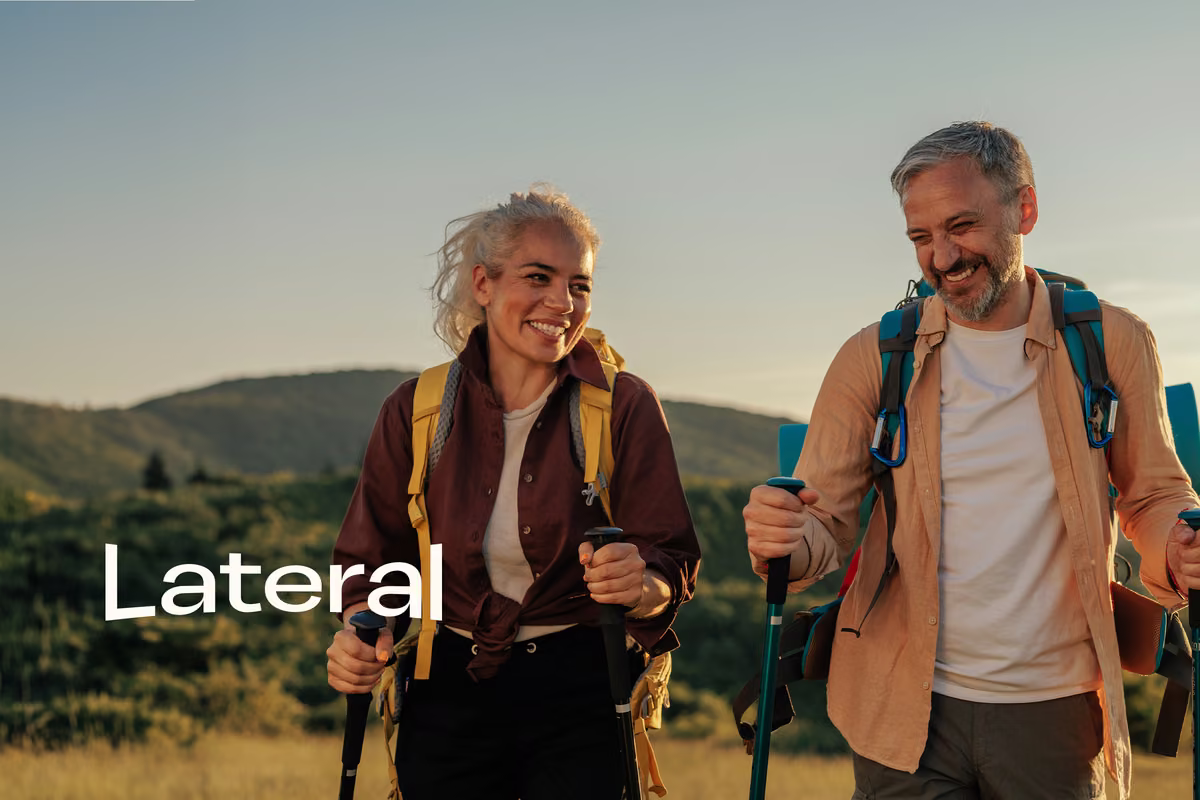 Two hikers smiling and walking outdoors with backpacks and trekking poles, green hills behind them, with the "Lateral" logo overlaid on the image.Two hikers smiling and walking outdoors with backpacks and trekking poles, green hills behind them, with the "Lateral" logo overlaid on the image.