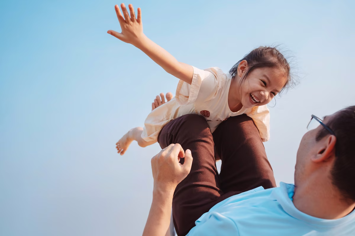 Father lifts laughing young girl overhead, arm outstretched, against a clear blue sky.