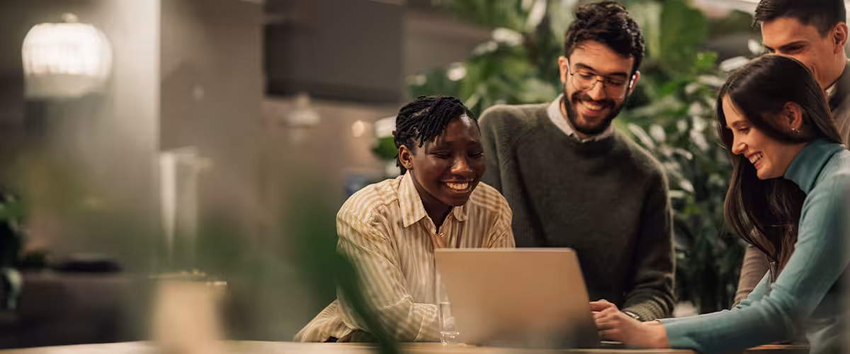 Four colleagues gather around a laptop in an office with lush green plants, smiling as they look at the screen together.