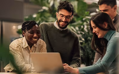 Four colleagues gather around a laptop in an office with lush green plants, smiling as they look at the screen together.