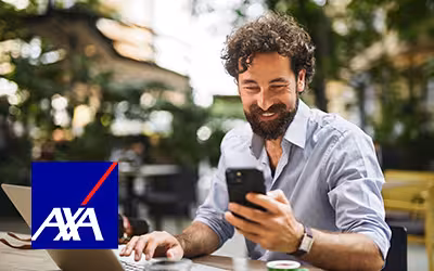 Man smiling while checking his phone at an outdoor café table, laptop open in front of him, with the AXA logo displayed in the corner."