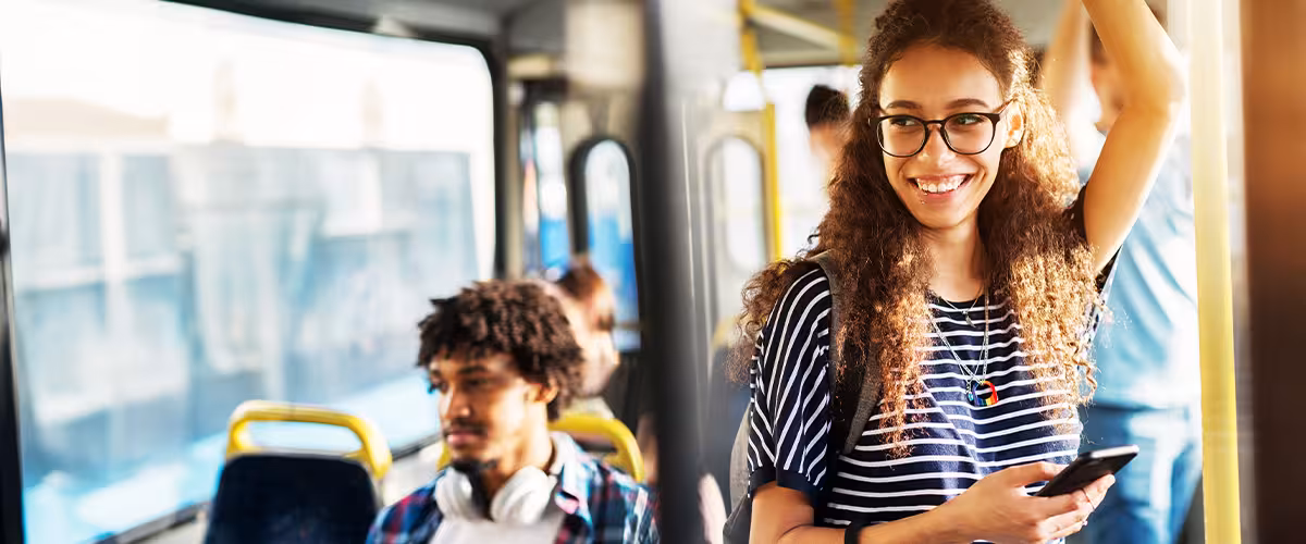 Young woman with curly hair and glasses smiling while standing on a bus, holding a phone and holding onto an overhead rail, with a backpack on her shoulder.