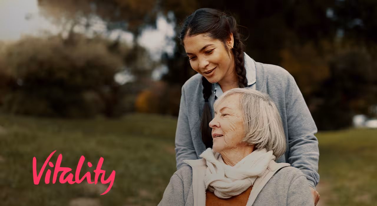 A younger woman smiling with an older woman outdoors, with the Vitality logo in the corner.