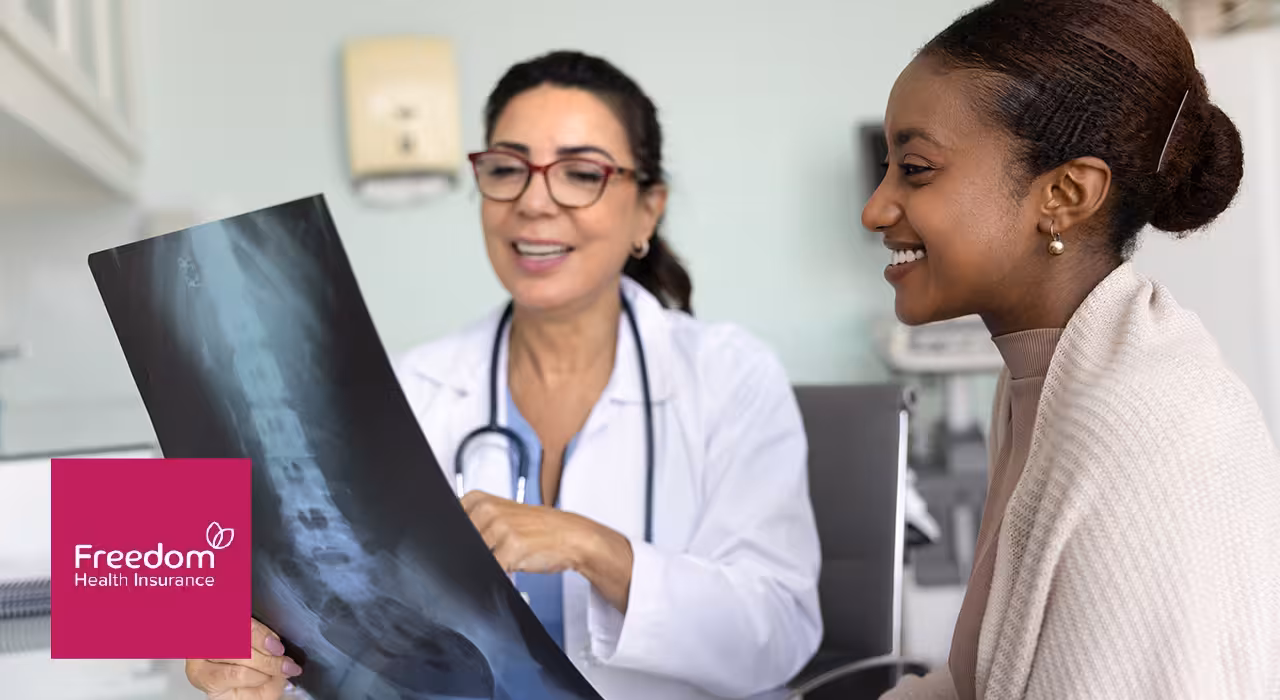 A doctor shows a smiling patient an X-ray of her spine during a consultation, with the Freedom Health Insurance logo in the corner.