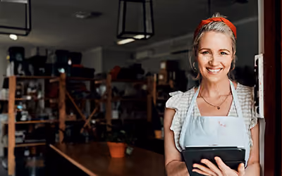 Smiling woman in an apron holding a tablet, standing in the doorway of a small shop or workshop with shelves of stock in the background