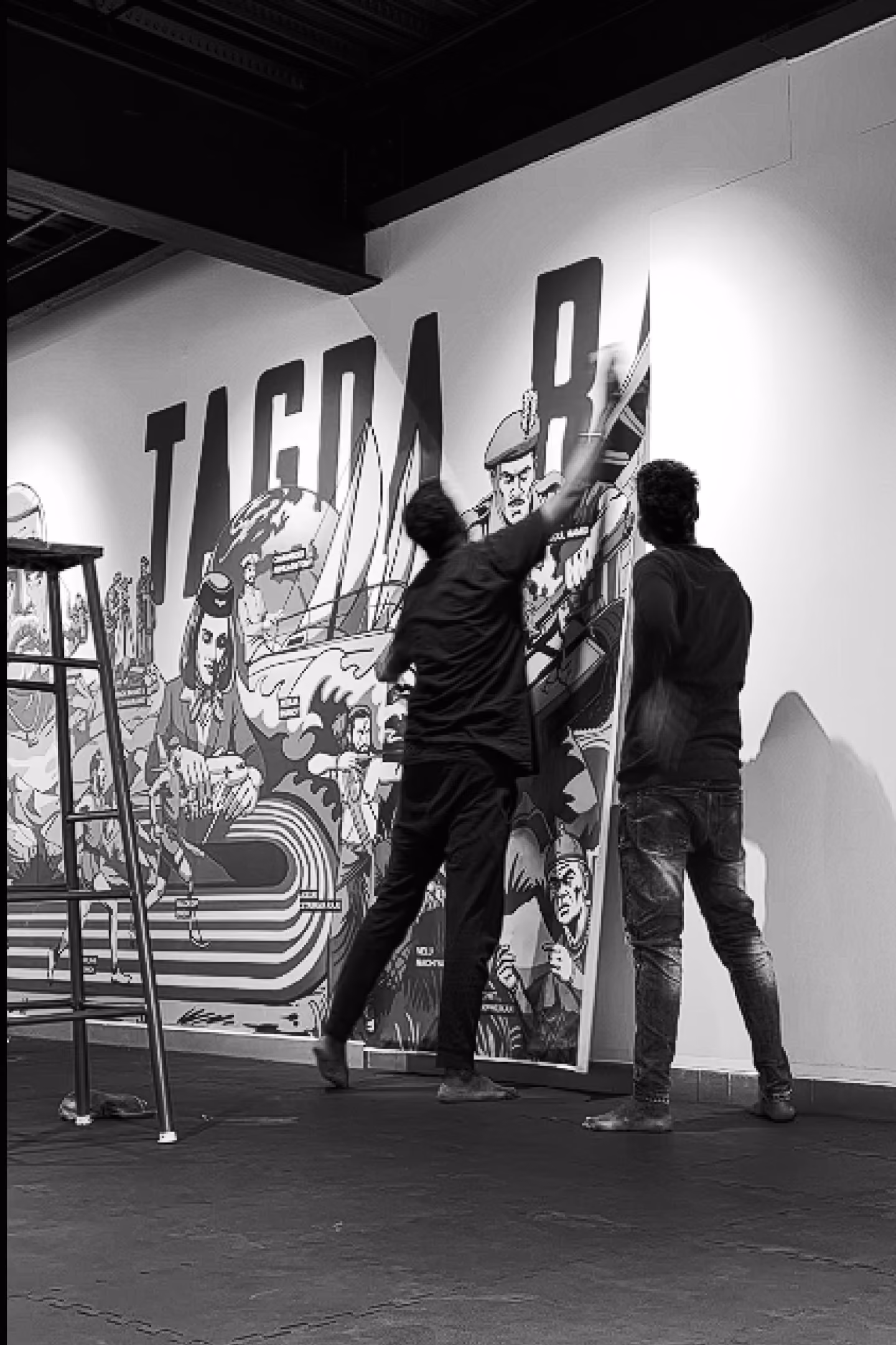 Two men installing or adjusting a large illustrated wall poster with the text 'TAGDA RAHO' and various artistic human and mechanical figures, in a room with a ladder and patterned floor mats.