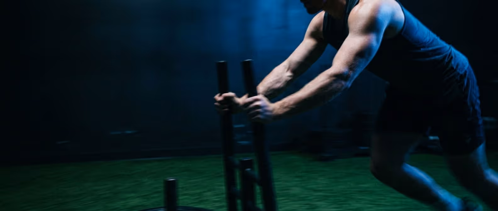 Muscular man pushing a weighted sled on artificial turf in a dimly lit gym.