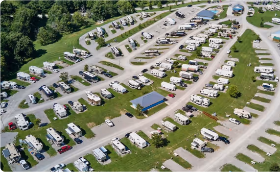 A large aerial view of an RV campground with rows of RVs and campers parked on green grass, crisscrossed by paved roads. Some areas feature small buildings and recreational spaces.