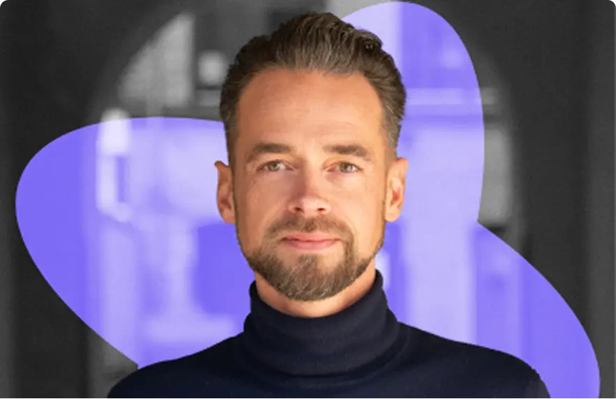 Man with short brown hair and beard wearing a black turtleneck, with a purple abstract shape in the background.