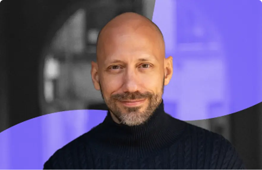 Bald man with a short beard wearing a black turtleneck sweater smiling against a blurred background with purple shapes.