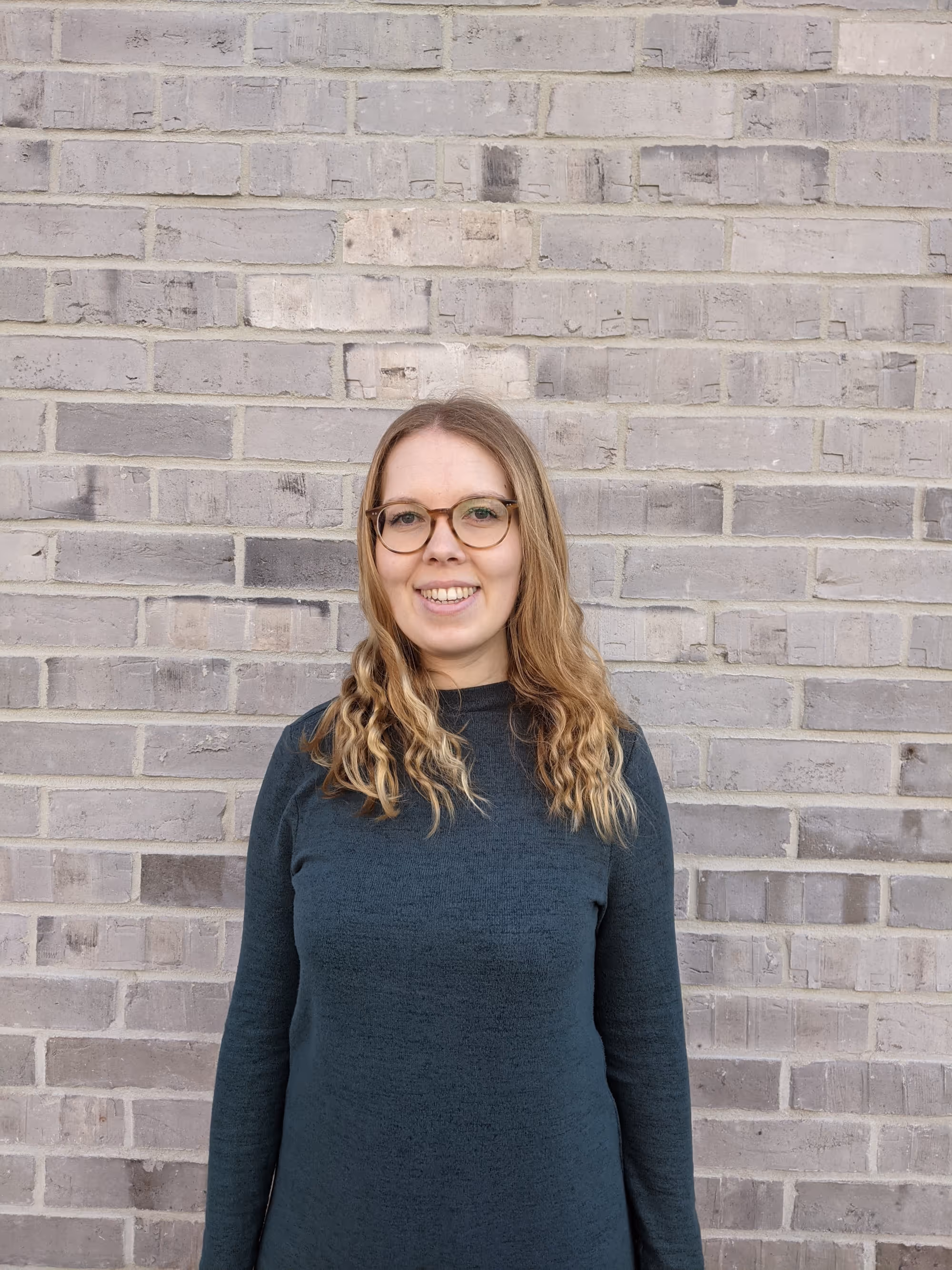 Smiling woman with glasses and wavy blonde hair wearing a dark green sweater stands against a gray brick wall.