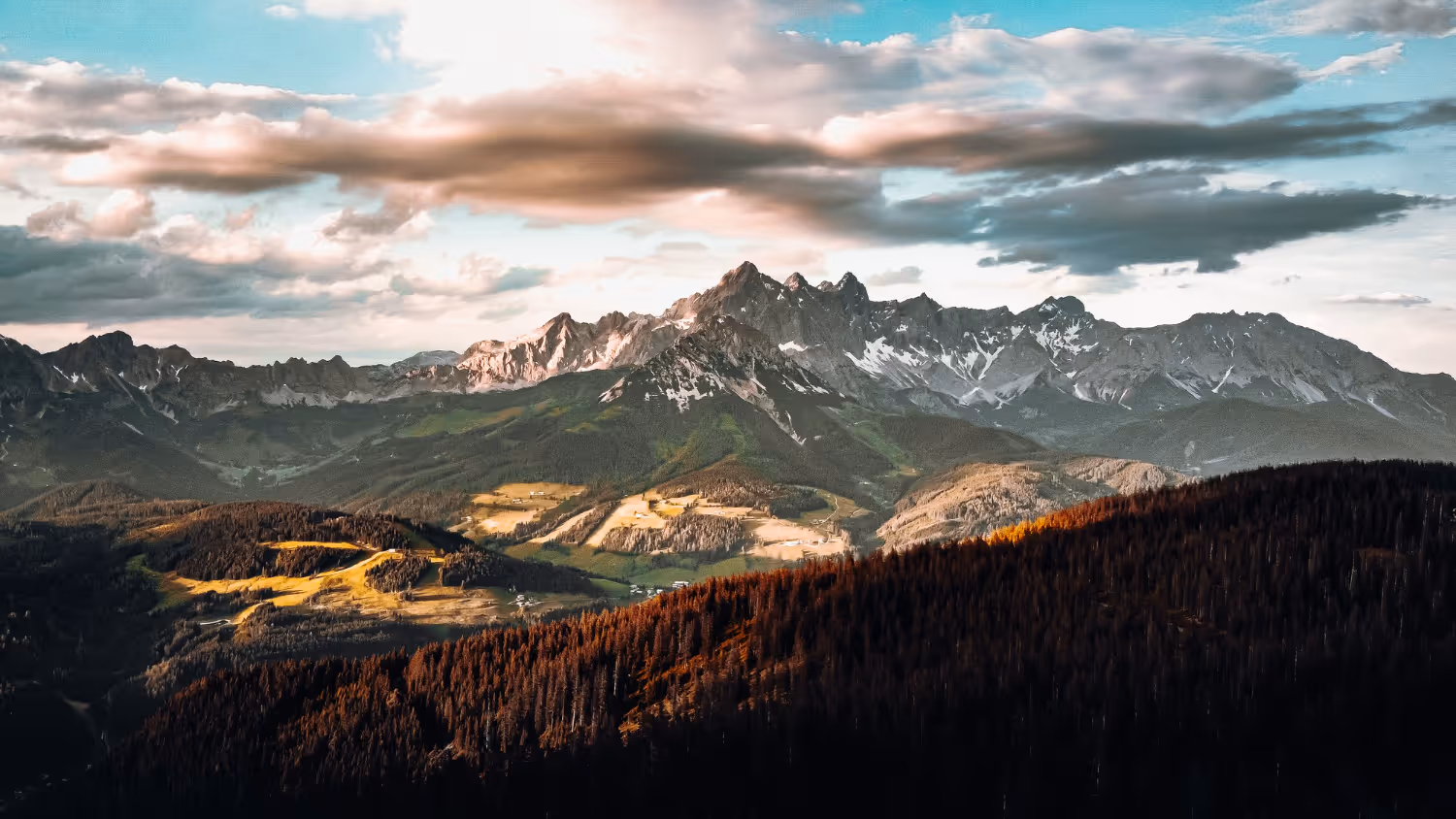 Mountain range with patches of snow and forested hills under a partly cloudy sky at sunset.
