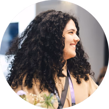 Smiling woman with curly black hair wearing a lanyard, looking to the right in a bright indoor setting.
