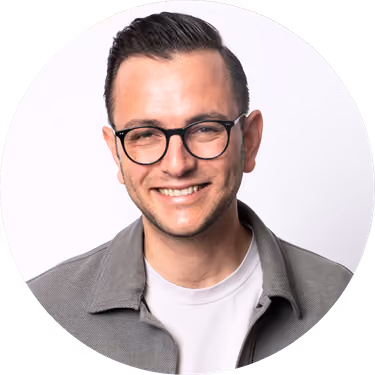 Smiling man with black glasses and dark hair wearing a gray jacket and white shirt on a white background.