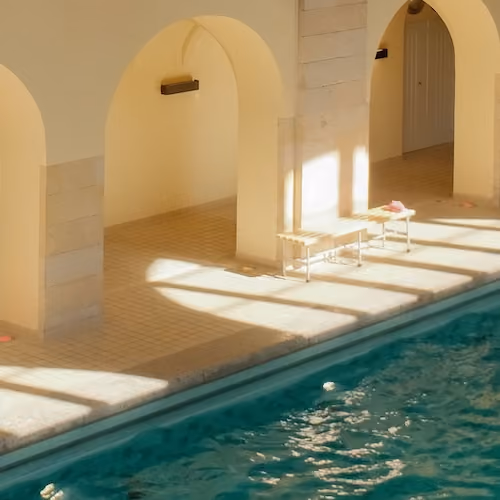 Indoor pool with sunlight casting shadows on tiled floor under arches and benches.