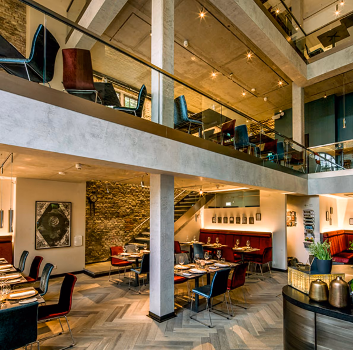 Modern two-level restaurant with stone walls, wooden floors, and mixed red and black chairs.