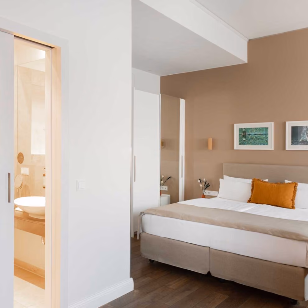 Modern bedroom with beige walls, a bed with white and brown linens, and a bathroom visible through an open door.