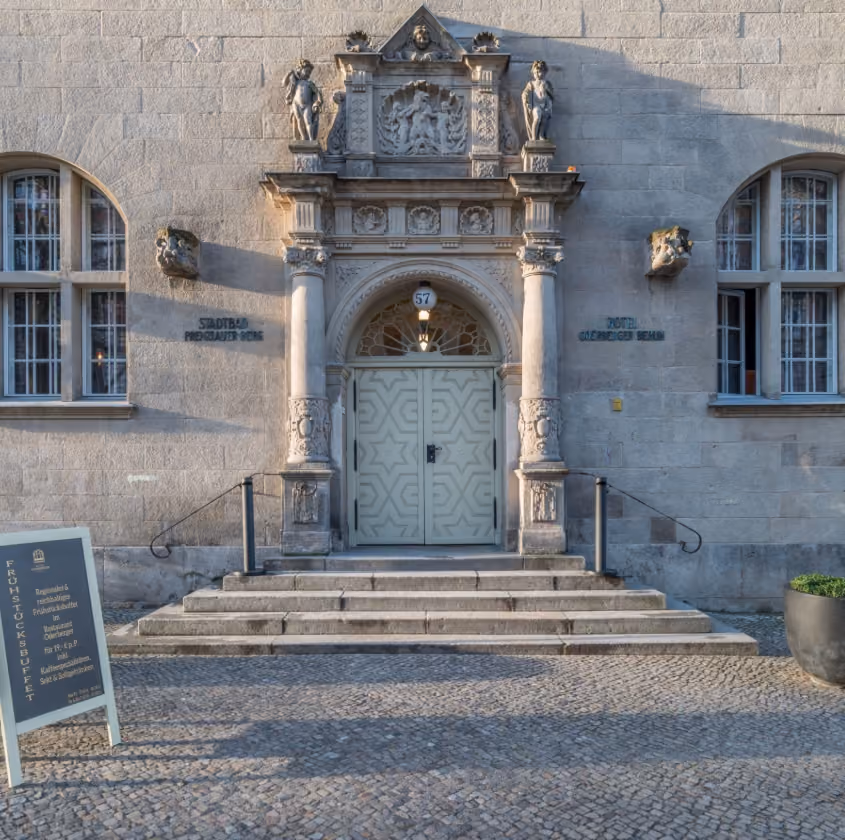 Ornate stone entrance with carved pillars and statues below two arched windows on a cobblestone street.