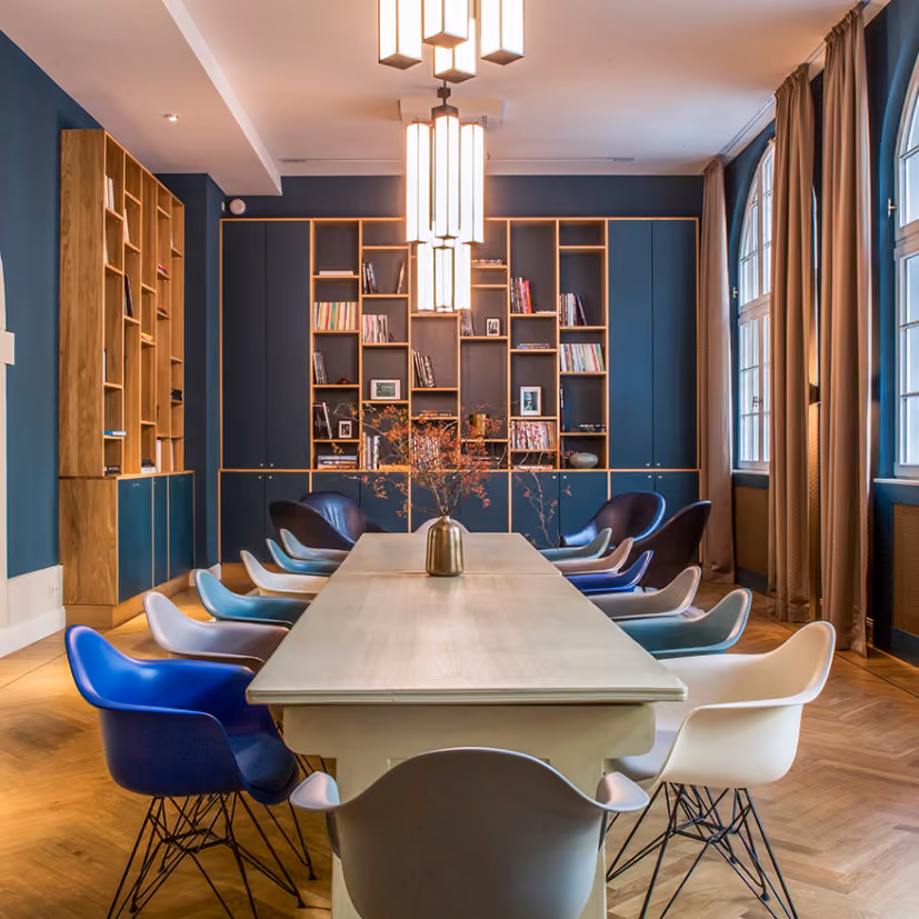 Long wooden table with mixed-color chairs in a room with tall bookshelves and arched windows.