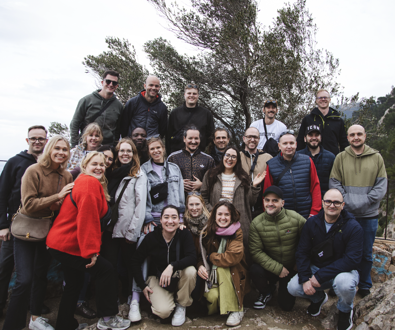 Group photo of 27 people outdoors with trees and mountains in the background on a cloudy day.