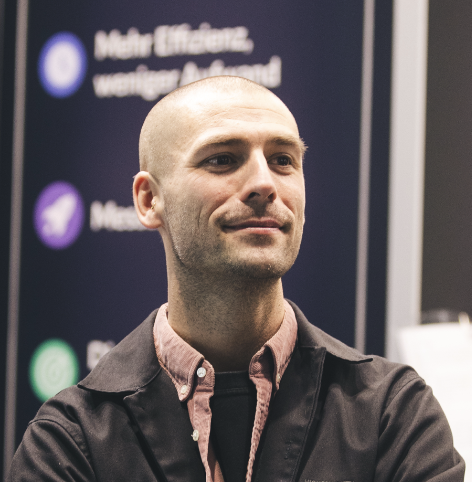 Smiling bald man in a black jacket and pink collar shirt stands against a blurred dark background.