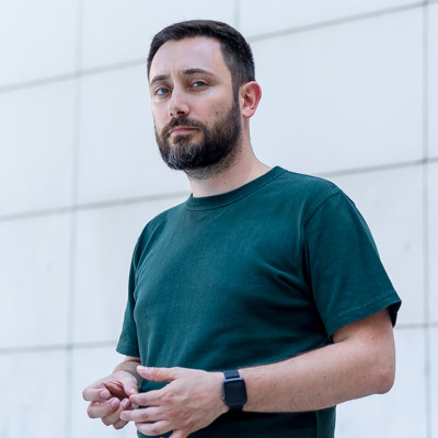 Bearded man in a dark green t-shirt and smartwatch looking at the camera against a white wall.