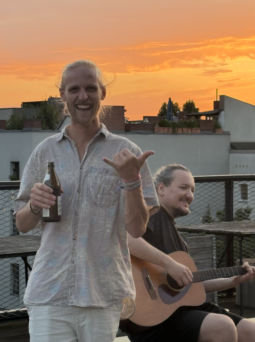 Man holding a beer bottle and making a shaka sign with a guitarist sitting behind him at sunset.