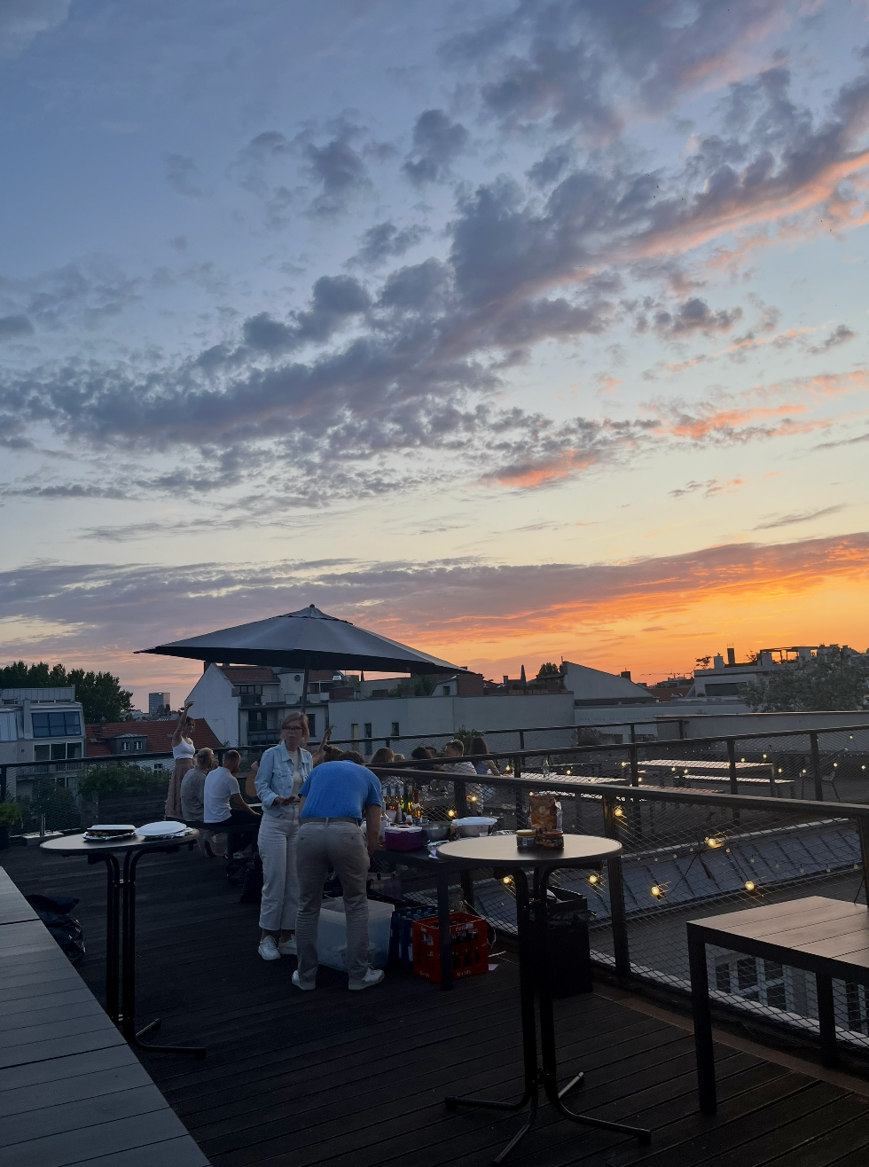People socializing on a rooftop deck at sunset with string lights and an umbrella.