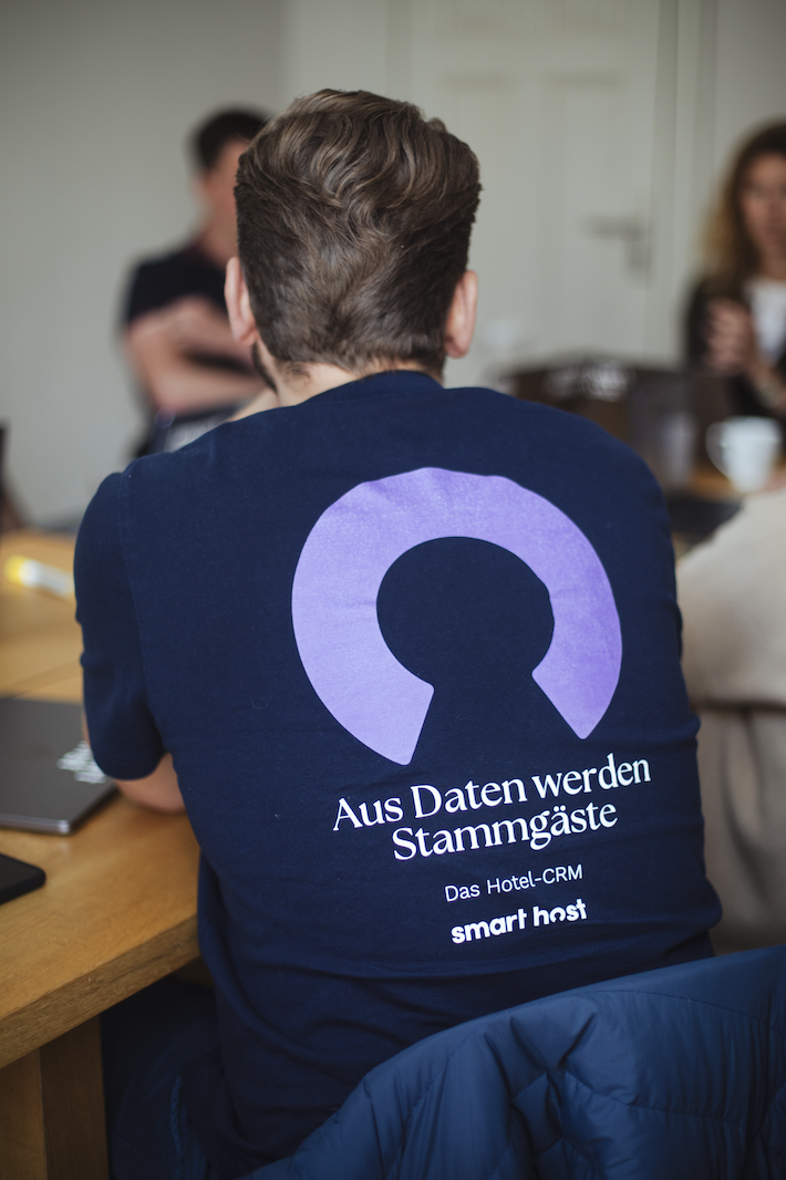 Man with short brown hair wearing a navy blue shirt with purple logo and German text on the back.