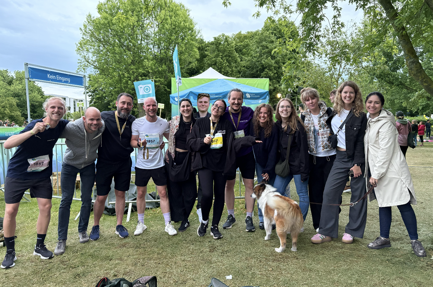 Group of 12 people smiling outdoors on grass under trees with a dog on a leash, some holding medals.