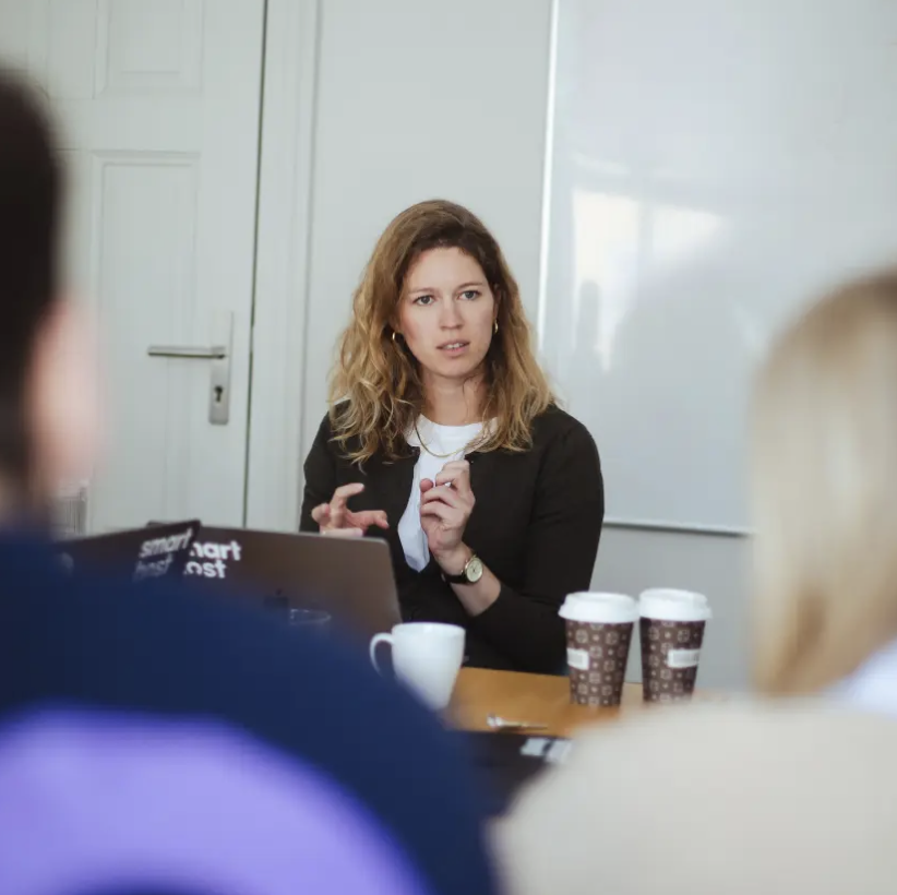 Woman with curly hair speaking in a meeting with two coffee cups and a laptop on the table.
