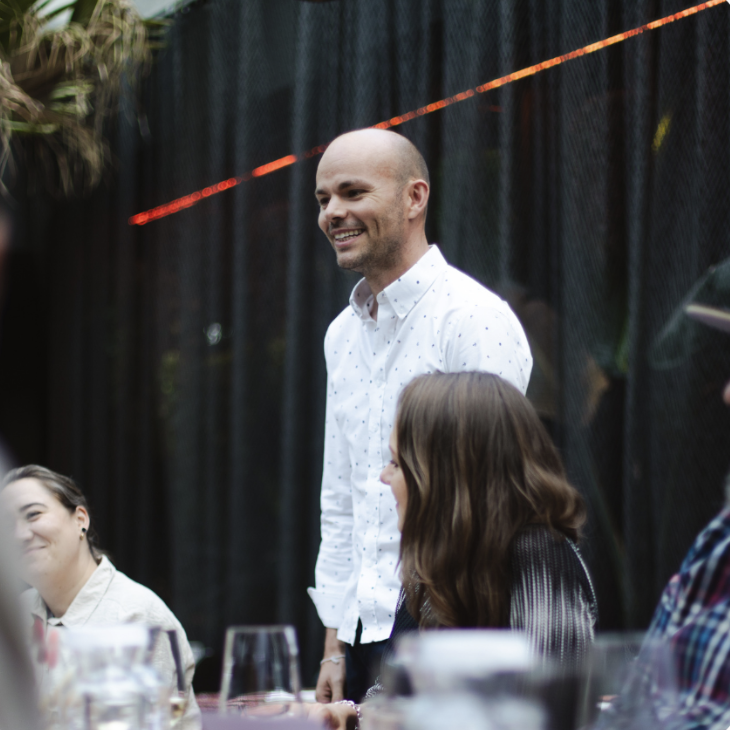 Smiling bald man in a white shirt standing and interacting with seated people around a table at an event.