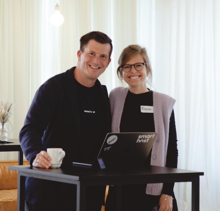 Two smiling people standing behind a black table with a laptop, one holding a white mug.