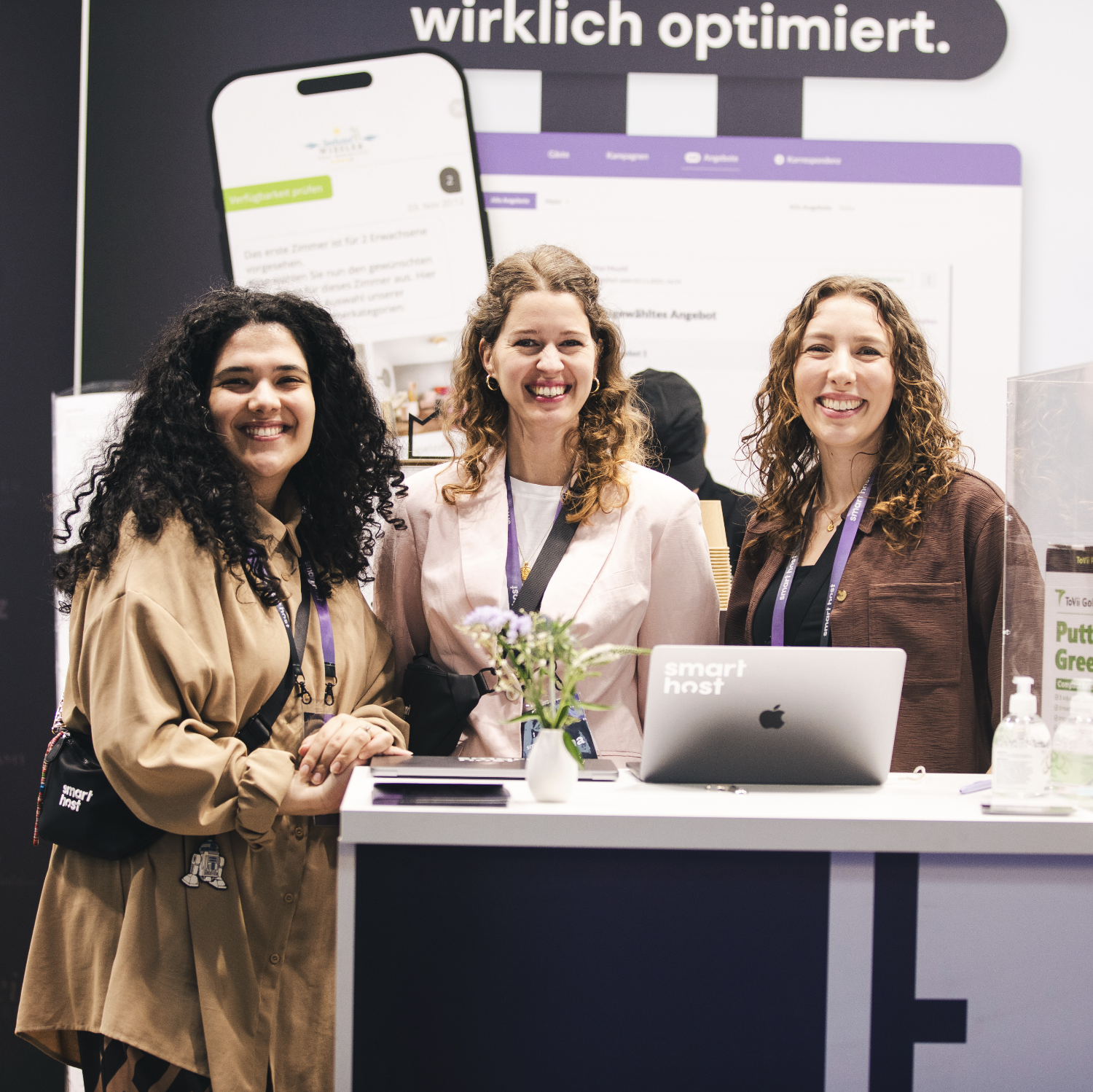 Three smiling women with curly hair stand behind a booth with a laptop that says smart host.