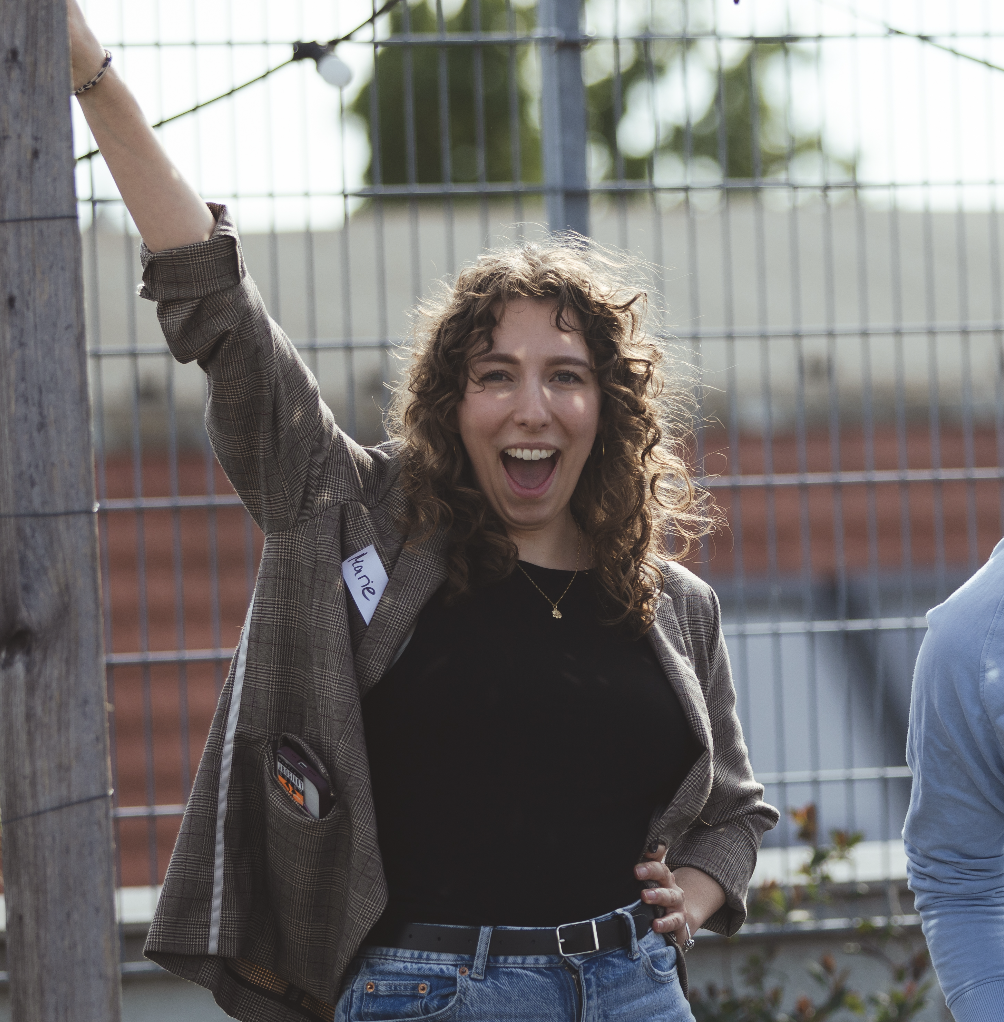 Woman with curly hair wearing a blazer and jeans, smiling with one arm raised outdoors.