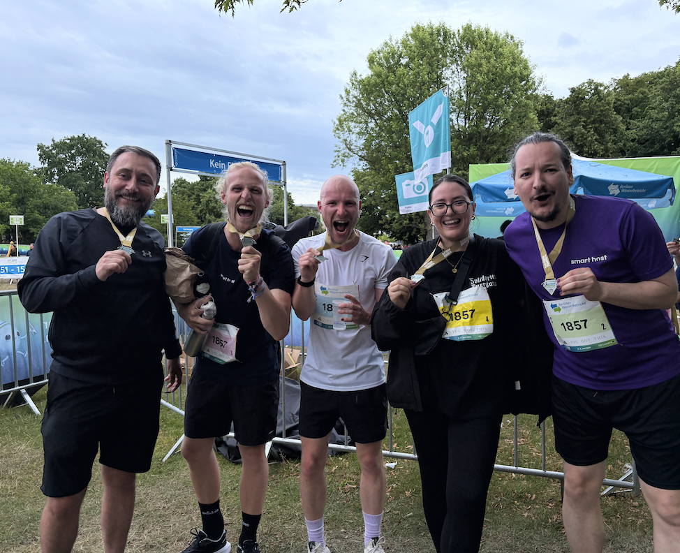 Five runners smiling and holding medals outdoors by a race finish area with greenery behind them.