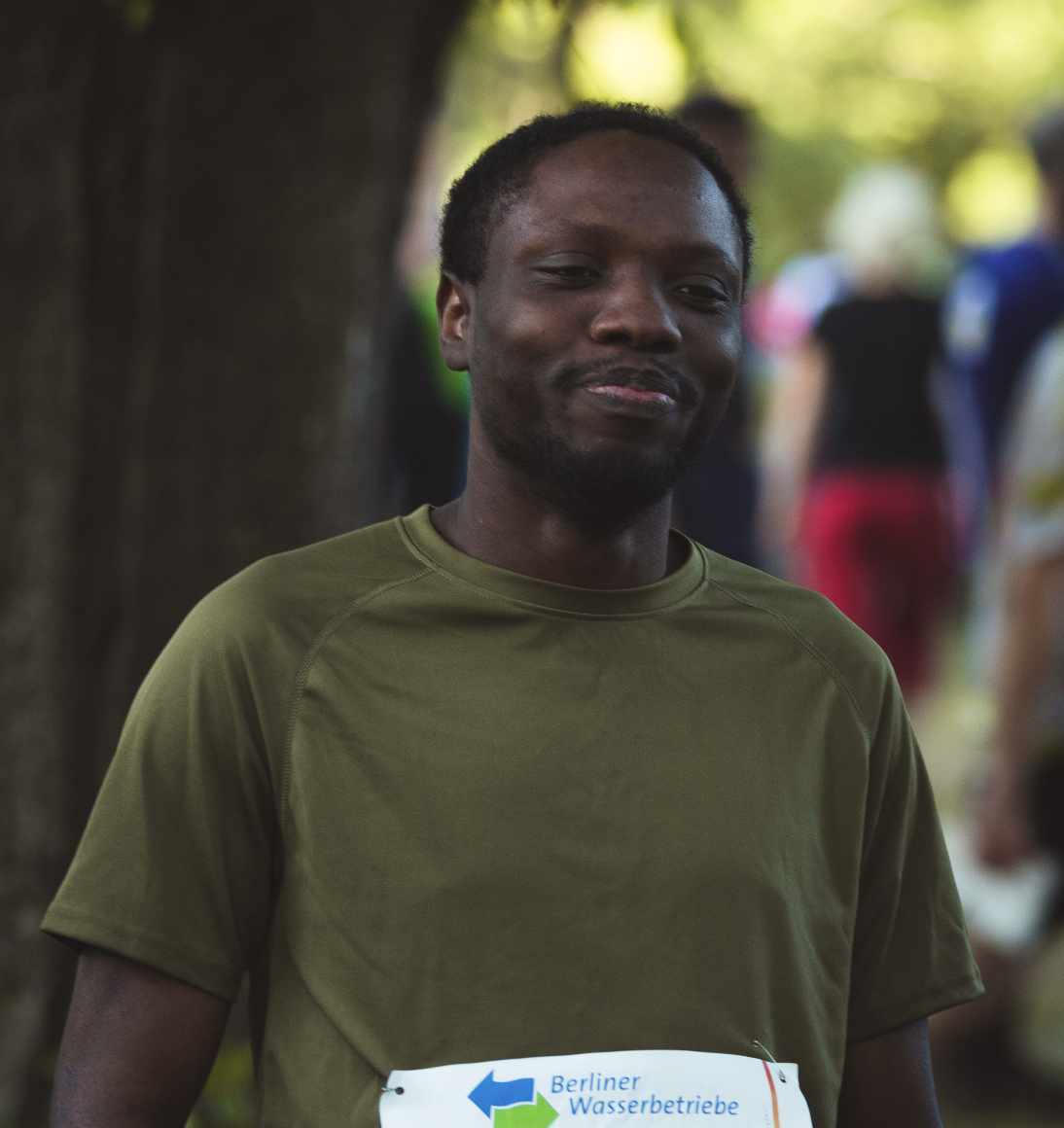 Man in olive shirt with Berliner Wasserbetriebe race bib outdoors, blurred people in background.