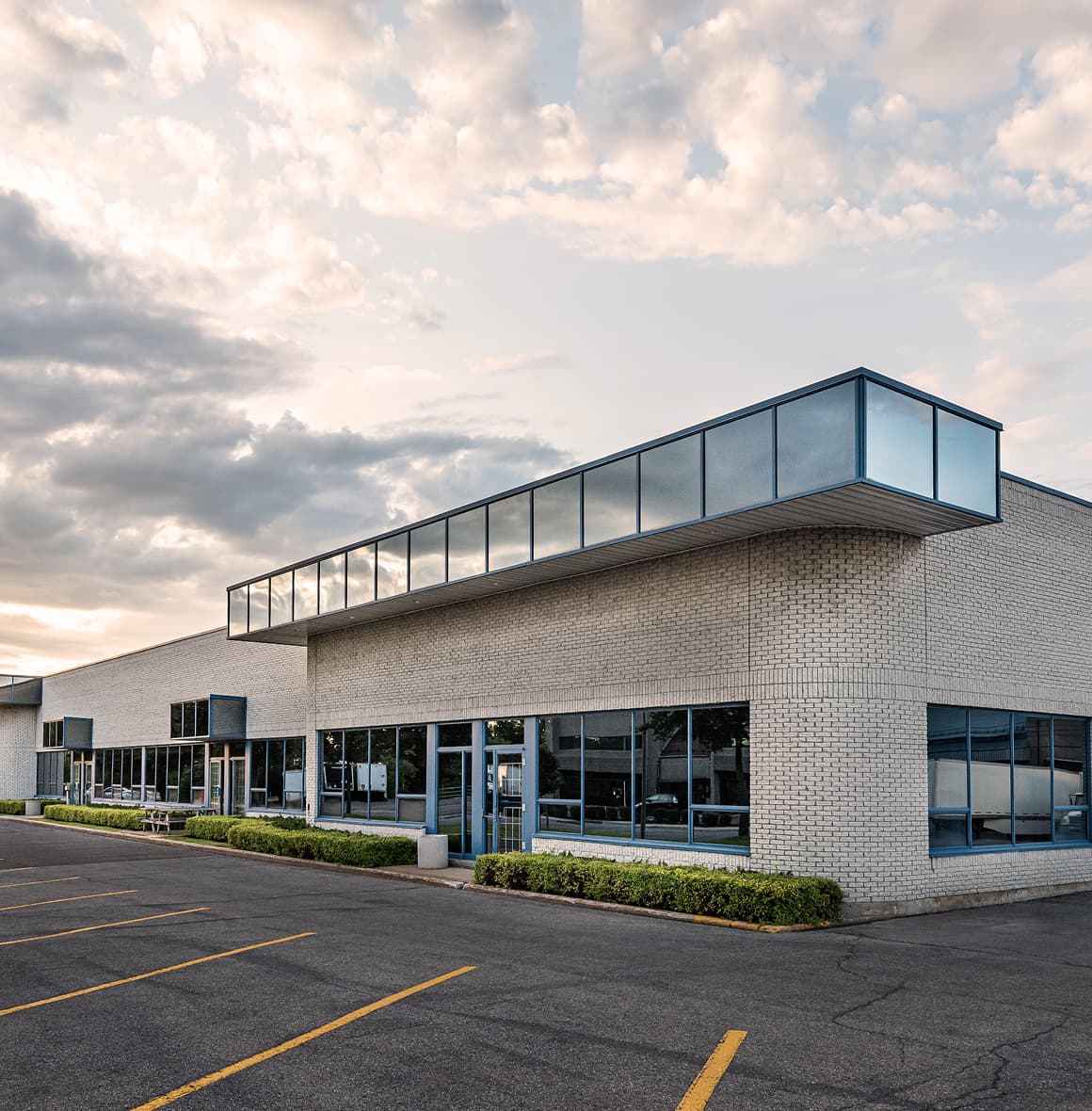A modern commercial or industrial building with white brick walls and large blue-tinted windows sits under a partly cloudy sky. The structure features a prominent glass overhang and an empty parking lot with yellow lines in the foreground