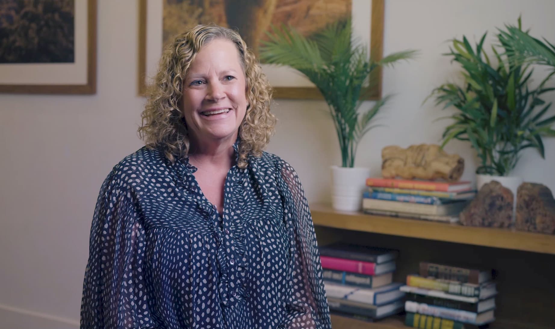 Smiling woman with curly blonde hair wearing a dark polka-dot blouse, seated in a cozy room with books, plants, and framed artwork in the background. The setting suggests a relaxed, home-office or interview environment.