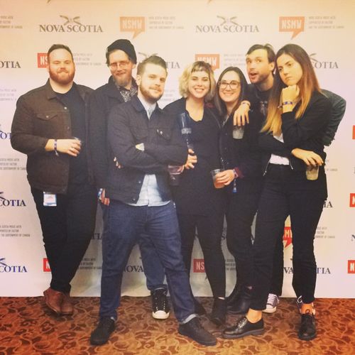 Group of seven people posing together in front of a step-and-repeat backdrop at an event, dressed in semi-casual attire, celebrating with drinks.