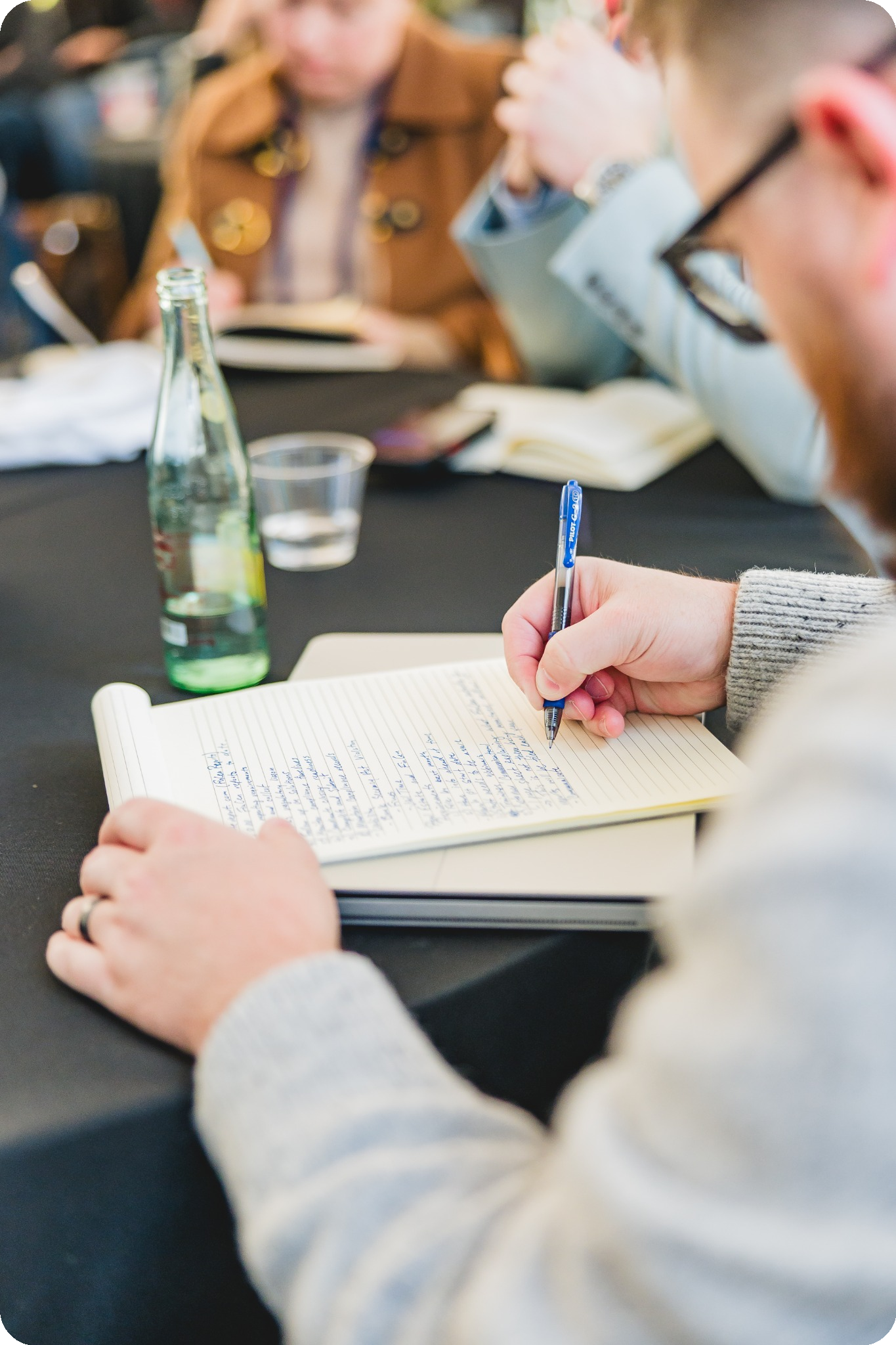 A person in a gray sweater writing notes on a notepad at a conference table. A bottle of sparkling water and a glass are nearby. The tone is focused and engaged.