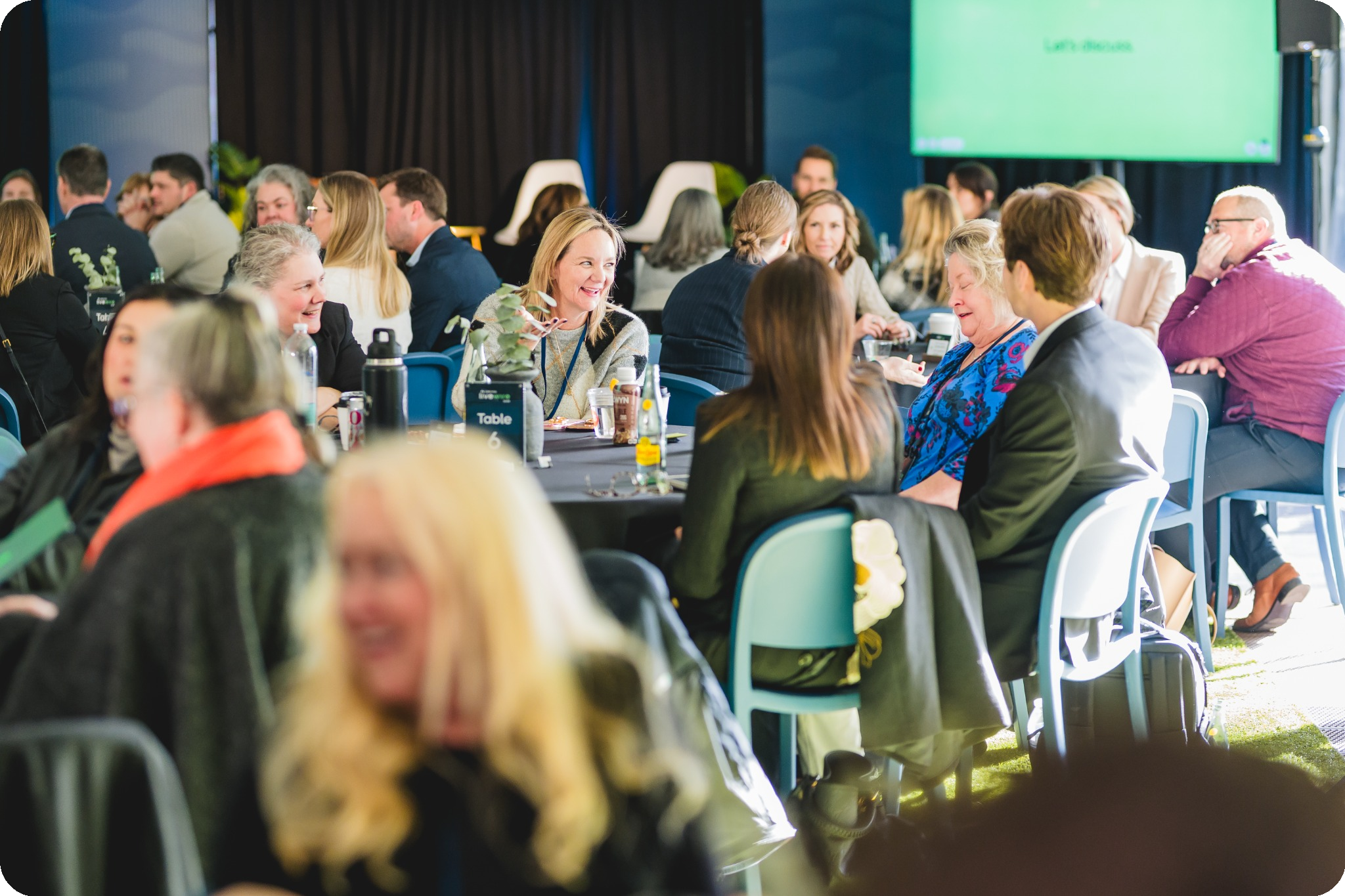 The room at LiveWire 2025 with diverse attendees in business attire seated at round tables, engaged in discussions and smiling.