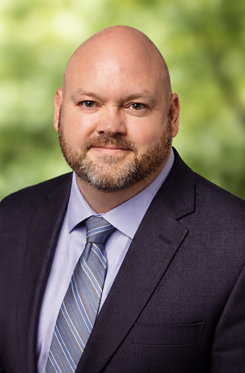 Portrait of Glenn Fredrickson wearing a dark suit, light blue shirt, and striped tie, with a blurred green background.