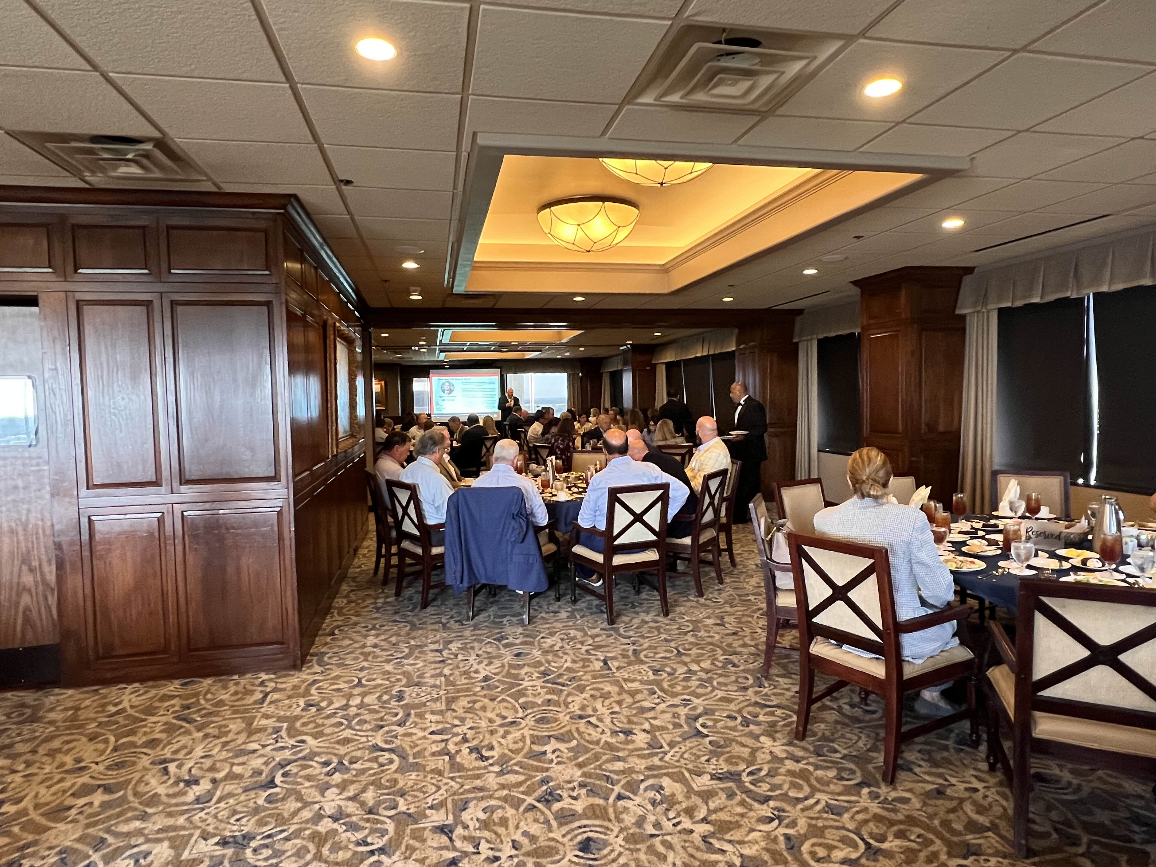 People seated at round tables in a carpeted, wood-paneled banquet room watching a speaker near a presentation screen.
