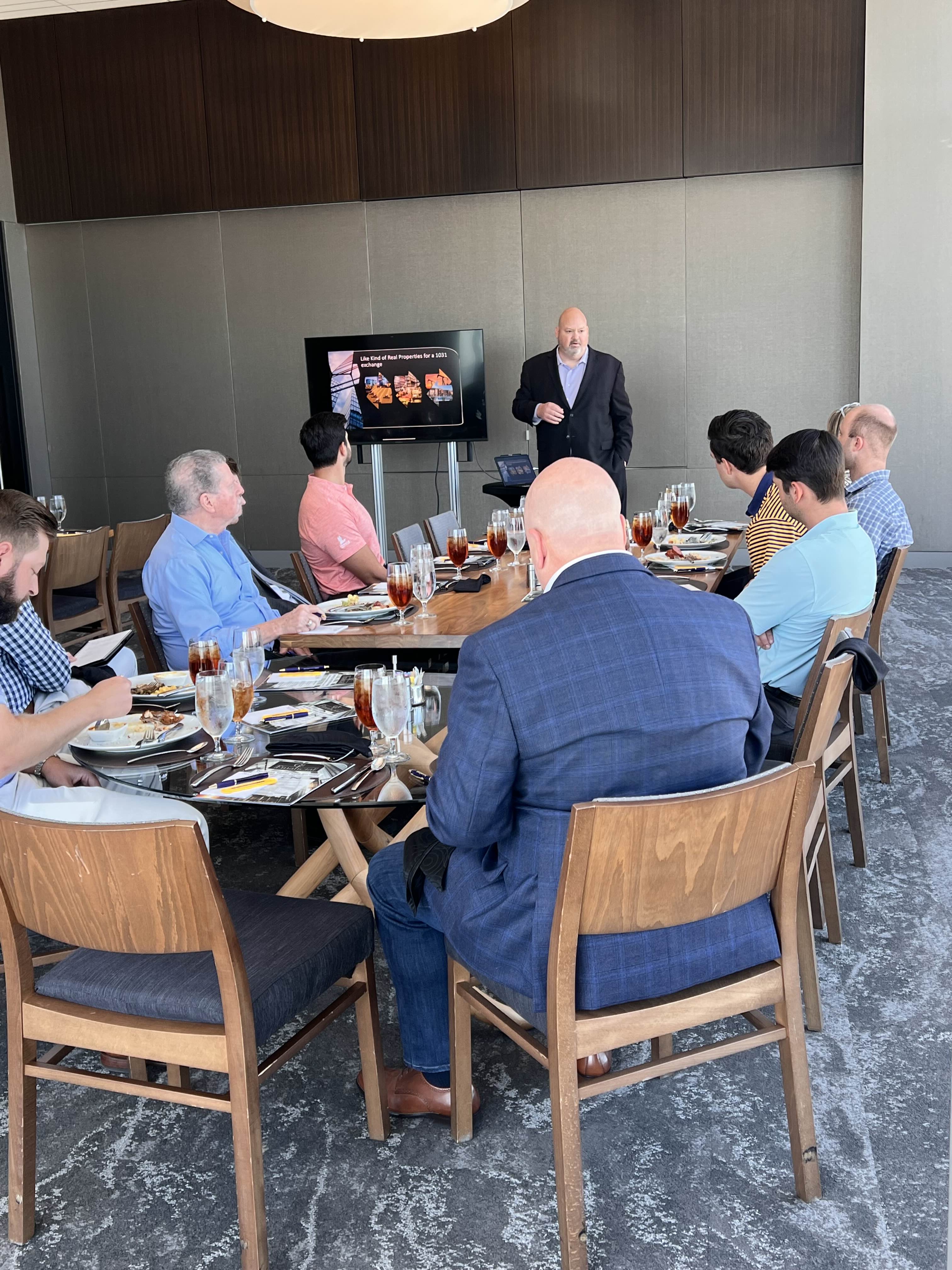 Business meeting with a presenter in a suit speaking to eight seated professionals around a large conference table with food and drinks.