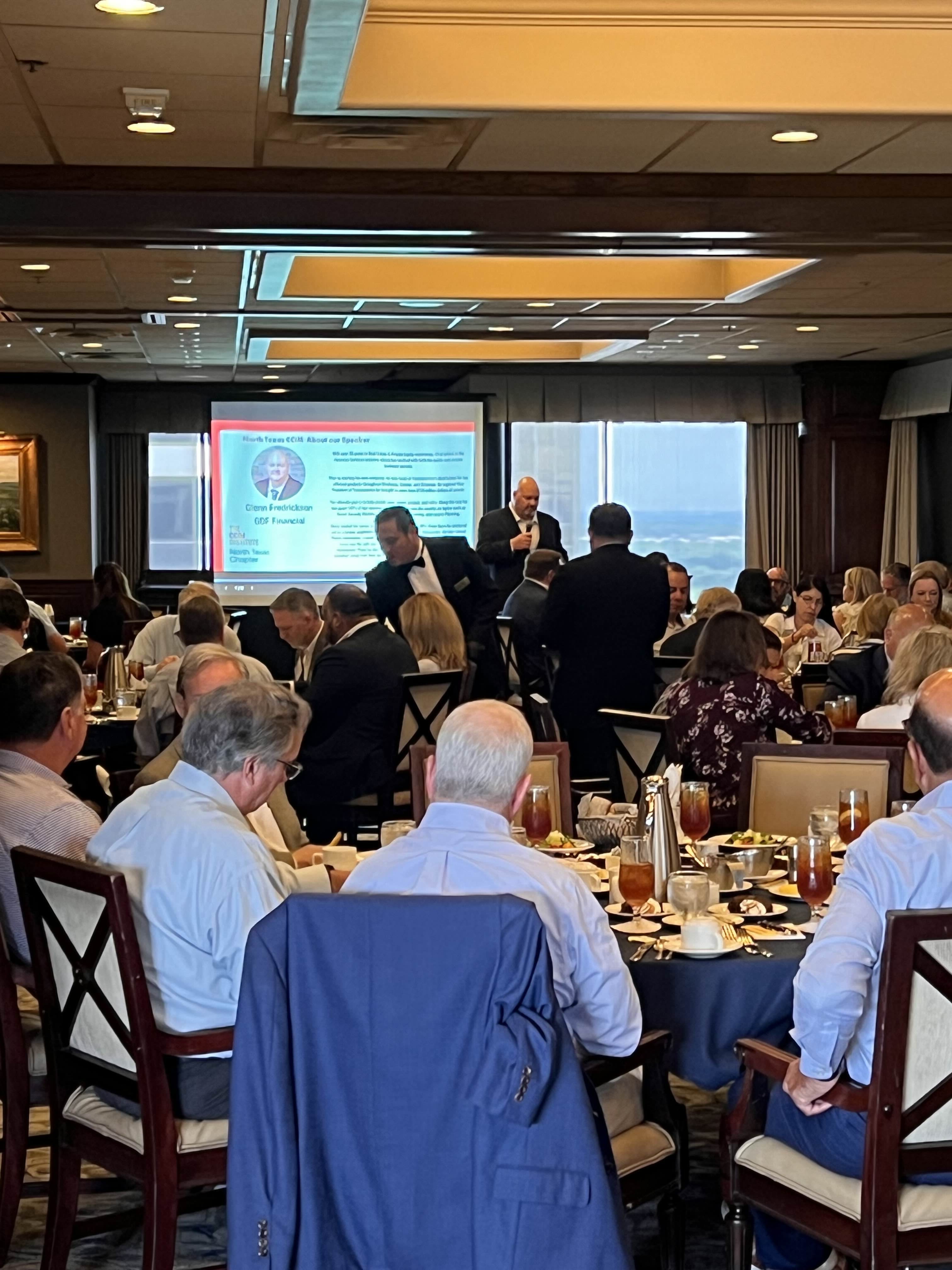 A formal luncheon event with attendees seated at round tables enjoying meals while a man speaks at the front near a presentation screen.
